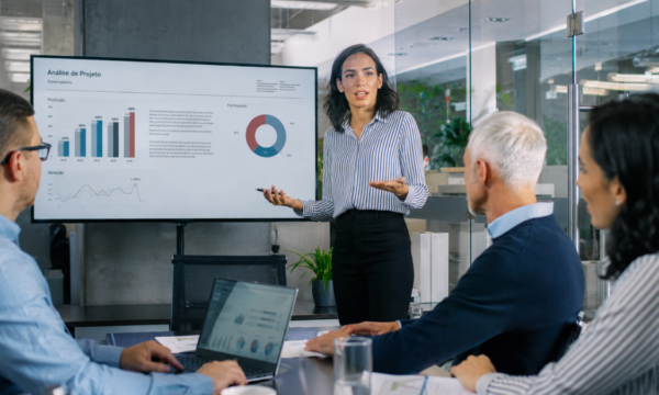 Profissional apresenta resultados e indicadores para uma equipe em reunião corporativa, representando a Trilha em Gestão de Projetos e a tomada de decisões baseada em dados.